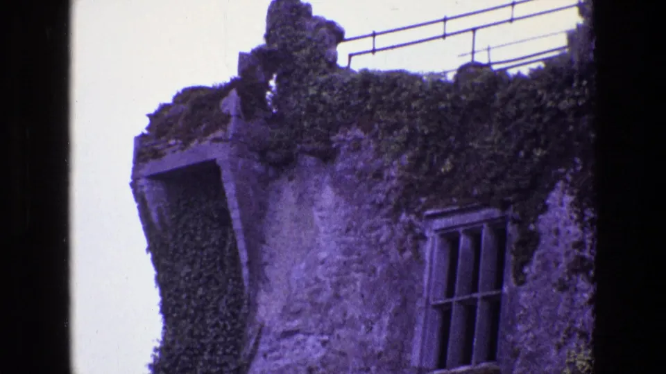 1969 Ireland 8mm frame showing a tilted visitor-shot view of a stone ruin corner with ivy, a barred window, and roof railing.