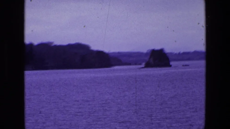 1969 Ireland 8mm frame showing calm water, a distant treeline, muted sky, and a small rock or island form offshore.