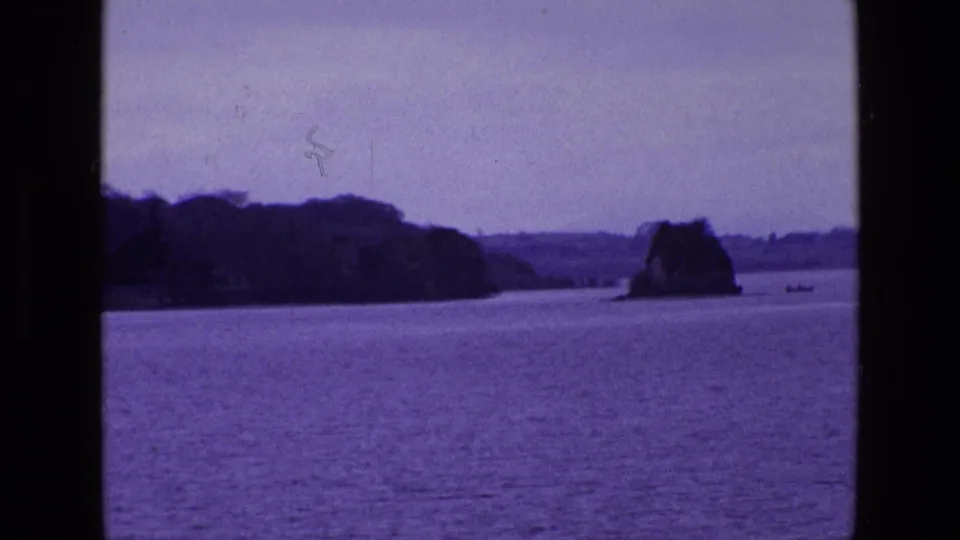 1969 8mm Ireland footage showing calm water, a rocky outcrop near shore, distant trees, pale sky, and a tiny boat on the horizon.