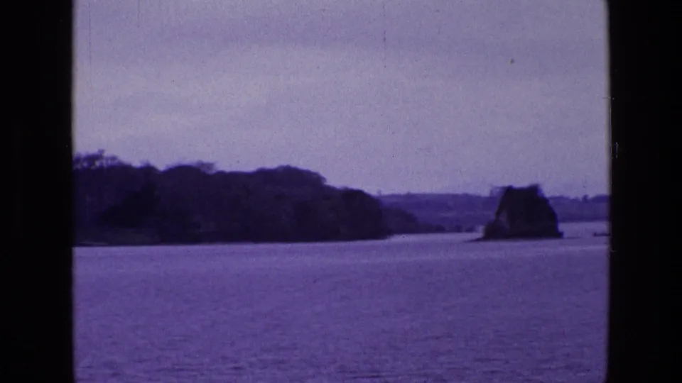 1969 Irish 8mm frame with open water in front, a dark tree-lined bank, rocky outcrop, and small boat in the distance.