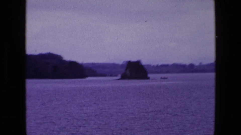Purple-shifted 1969 8mm Ireland scene with calm water, wooded shoreline, low distant hills, rocky outcrop, and a small boat.