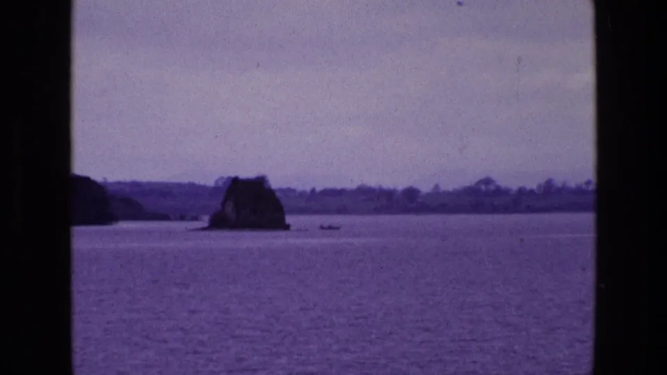 Late-1960s Irish 8mm frame centering a rocky outcrop across calm water, with a distant shoreline, pale sky, and tiny boat nearby.