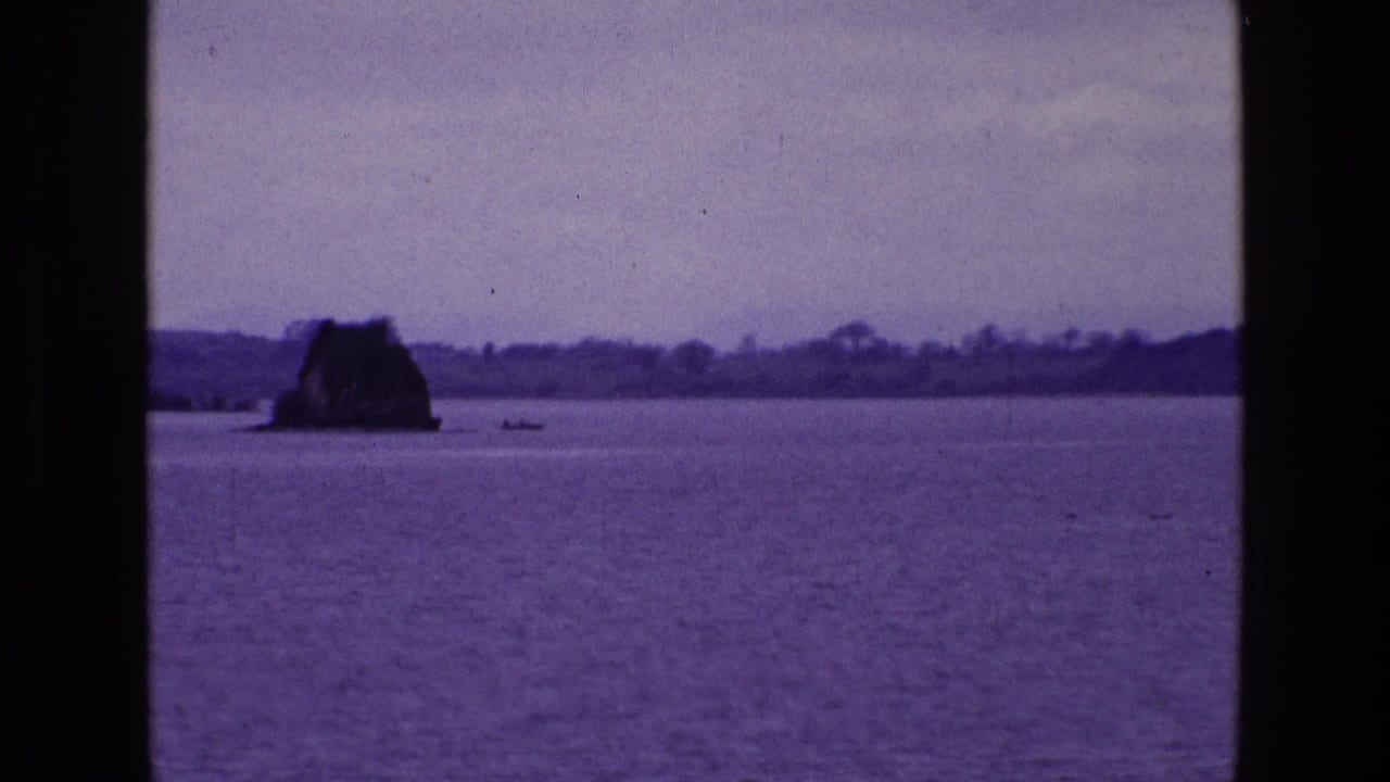 1969 8mm home-movie view in Ireland showing a lake, small rocky island, distant shoreline, dark window framing, and a tiny boat on the water.