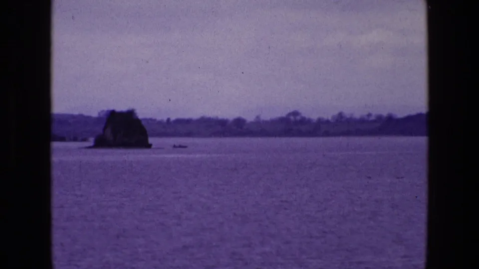 1969 8mm home-movie view in Ireland showing a lake, small rocky island, distant shoreline, dark window framing, and a tiny boat on the water.