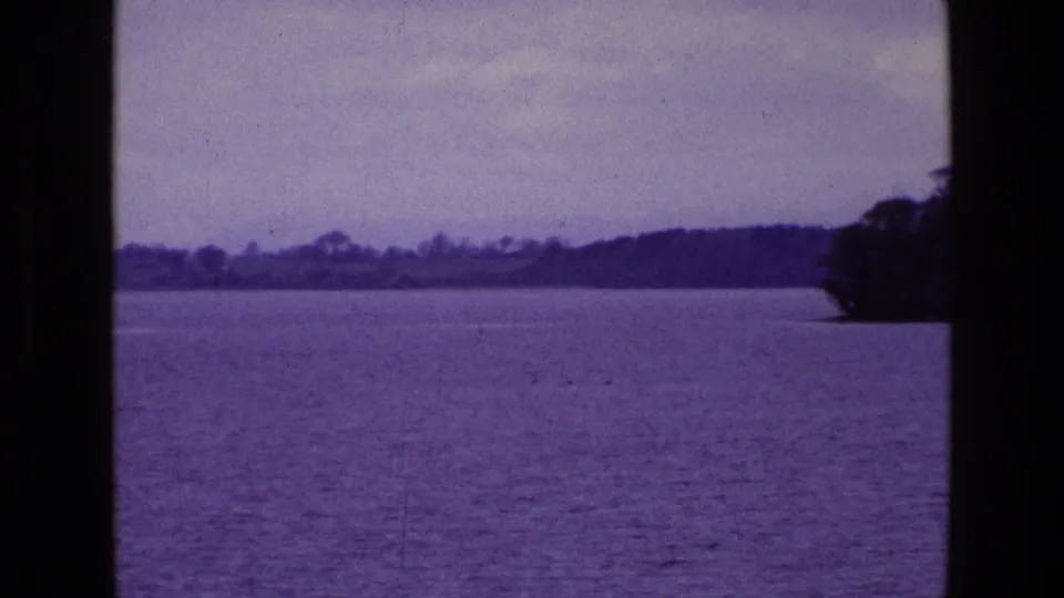 Soft purple 8mm frame of an Irish lake, distant wooded shoreline, pale sky, and dark window framing, with land entering at the right edge.