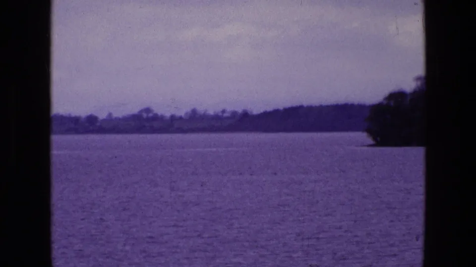 1969 Ireland home-movie frame showing a broad lake, distant shoreline, pale overcast sky, dark window bars, and trees along the right edge.
