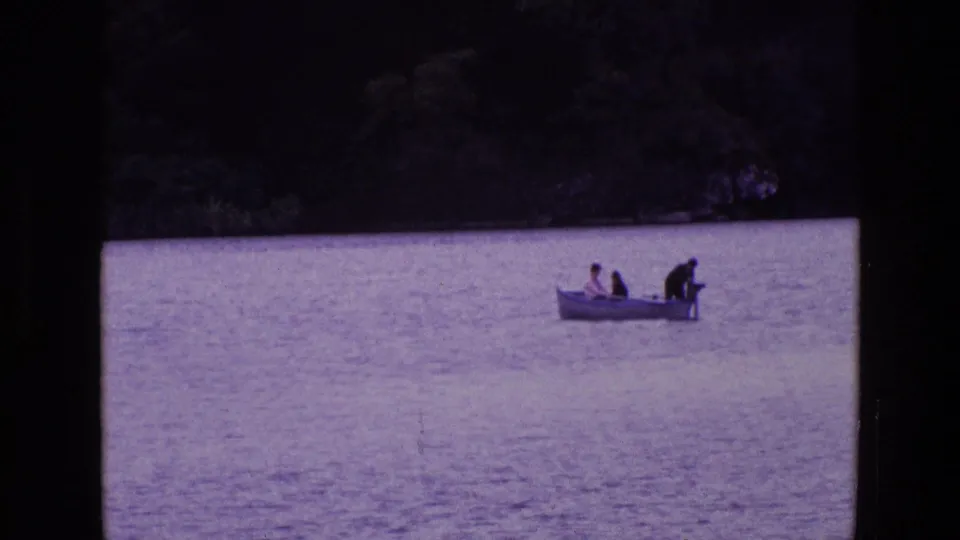1969 Ireland home-movie frame of a small motorboat crossing a lake, with two seated passengers and one person at the stern.