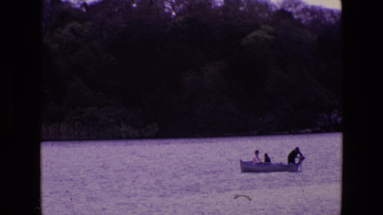1969 Ireland 8mm frame showing a small white rowboat with three distant passengers on calm lake water near trees and reeds.