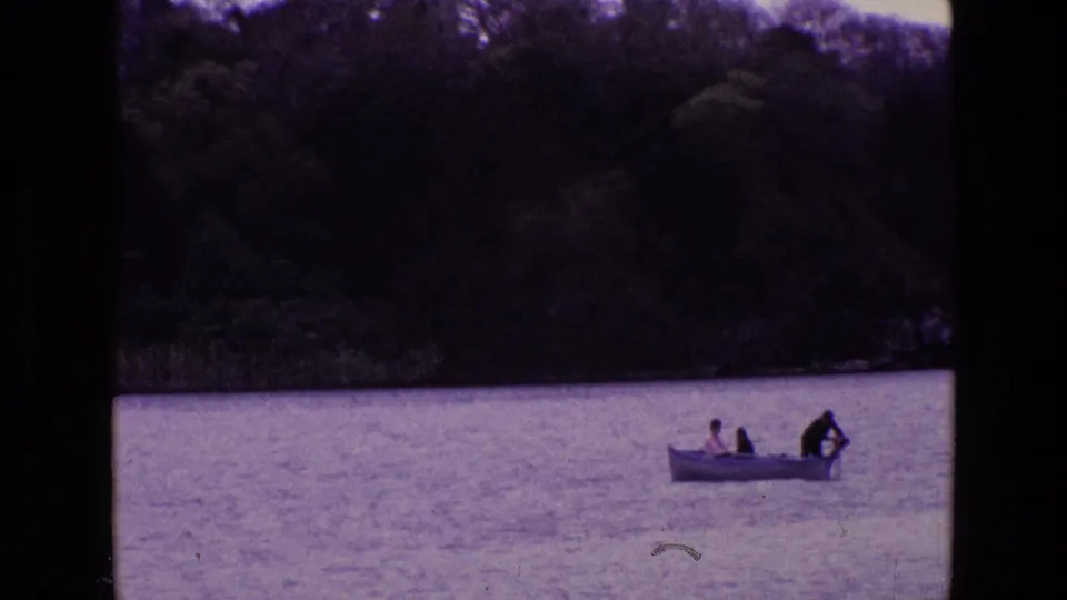 1969 Ireland 8mm frame showing a small white rowboat with three distant passengers on calm lake water near trees and reeds.