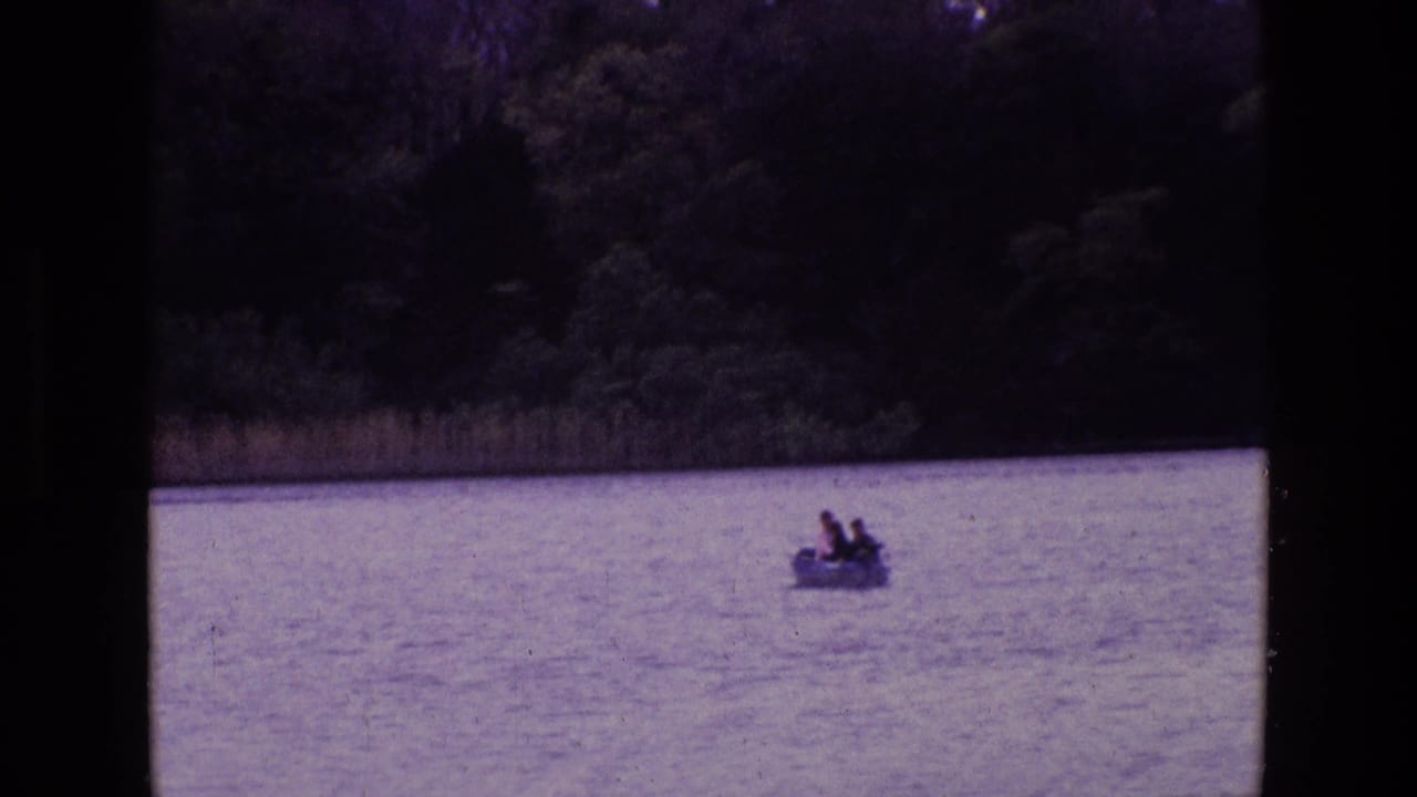 1969 Ireland home-movie frame with a small rowboat and distant passengers on open lake water against a wooded shore.
