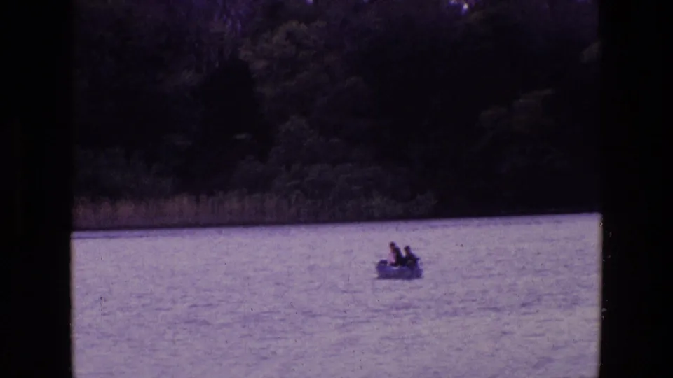 1969 Ireland home-movie frame with a small rowboat and distant passengers on open lake water against a wooded shore.