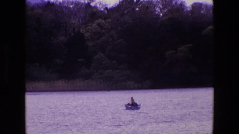 Distant 1969 Ireland 8mm view of a rowboat on a lake, framed below a dark wooded shoreline and visible film borders.