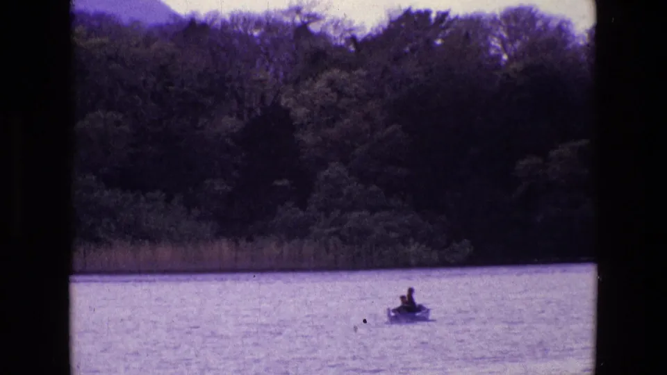 1969 Ireland home-movie still of a small rowboat with distant people on bright lake water near a wooded shoreline.