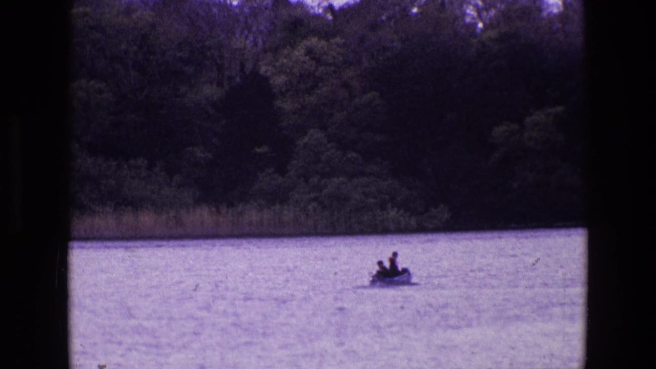 1969 Ireland 8mm frame showing two distant people in a small boat on calm water with trees and reeds along the shore.