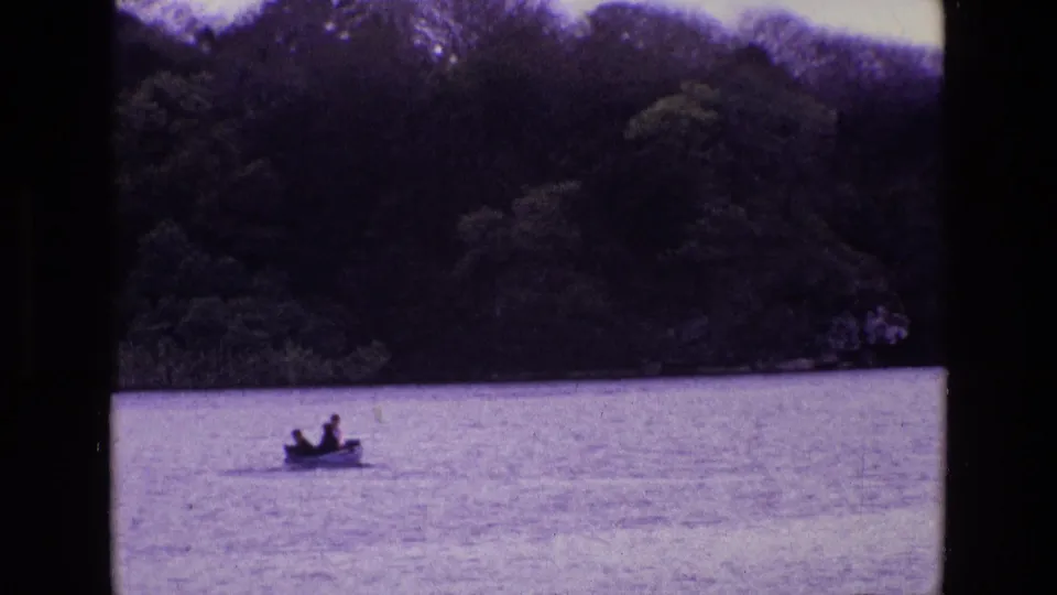1969 Ireland home-movie frame with a small open boat and two distant figures on a lake or broad waterway beneath trees.