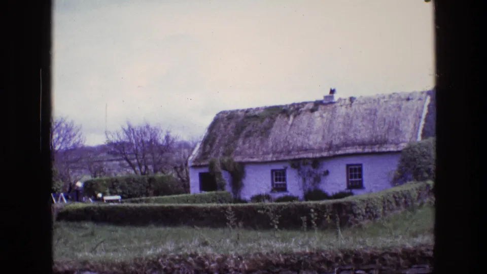 Rural Irish cottage exterior with thatched roof and hedges, seen in faded 1969 8mm home-movie color.