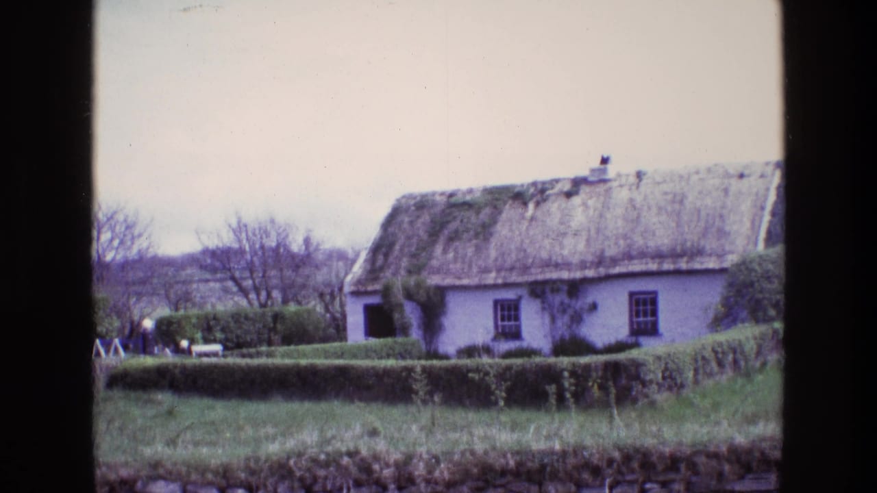 1969 Ireland 8mm view of a white thatched cottage with a trimmed hedge, bare trees, grass, and a dark vertical foreground edge.