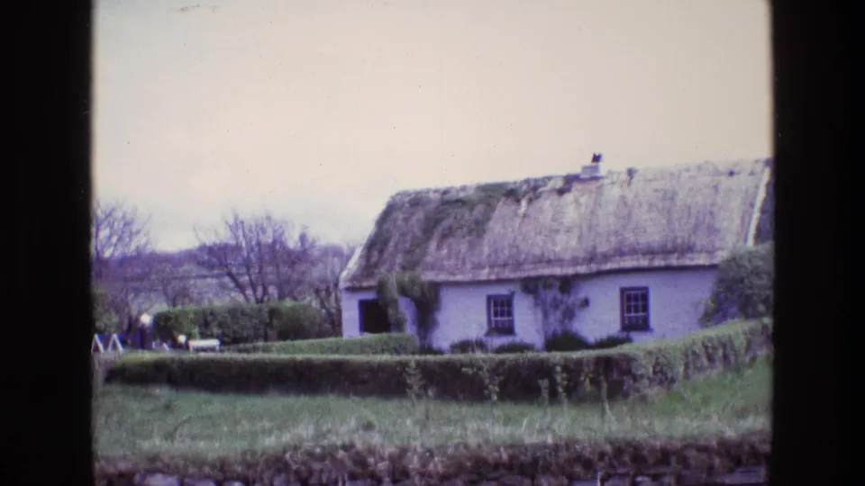 1969 Ireland 8mm view of a white thatched cottage with a trimmed hedge, bare trees, grass, and a dark vertical foreground edge.
