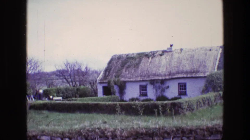 1969 Ireland 8mm still showing a thatched white cottage set behind a green hedge, with a grassy foreground and trees behind.