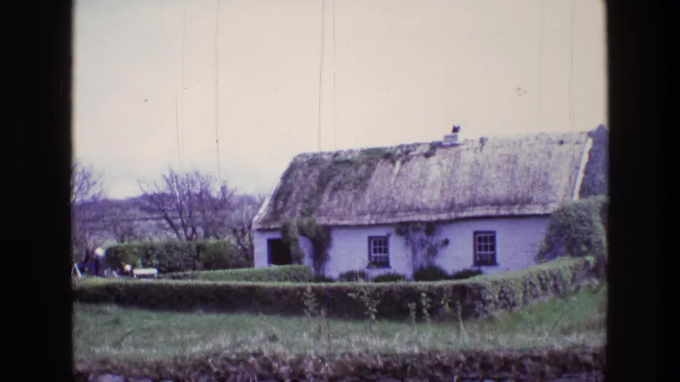 1969 Irish countryside frame showing a thatched cottage behind a hedge, with ivy, bare trees, low hills, and distant people near the left side.