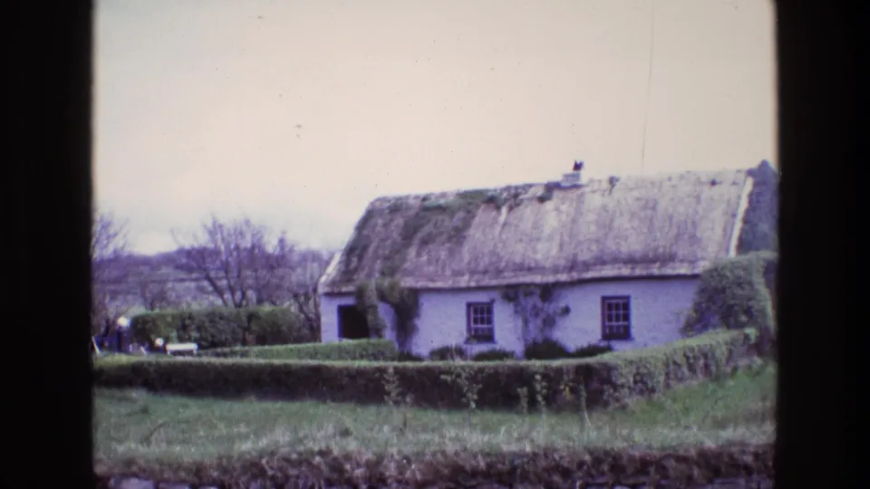 Final 8mm frame of the Irish rural cottage exterior, showing thatch, white walls, garden hedge, stone wall, grass, trees, and distant figures.
