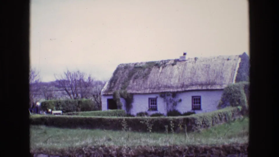 1969 Ireland 8mm frame showing a whitewashed thatched cottage behind hedges, grass, bare trees, and a low stone wall.