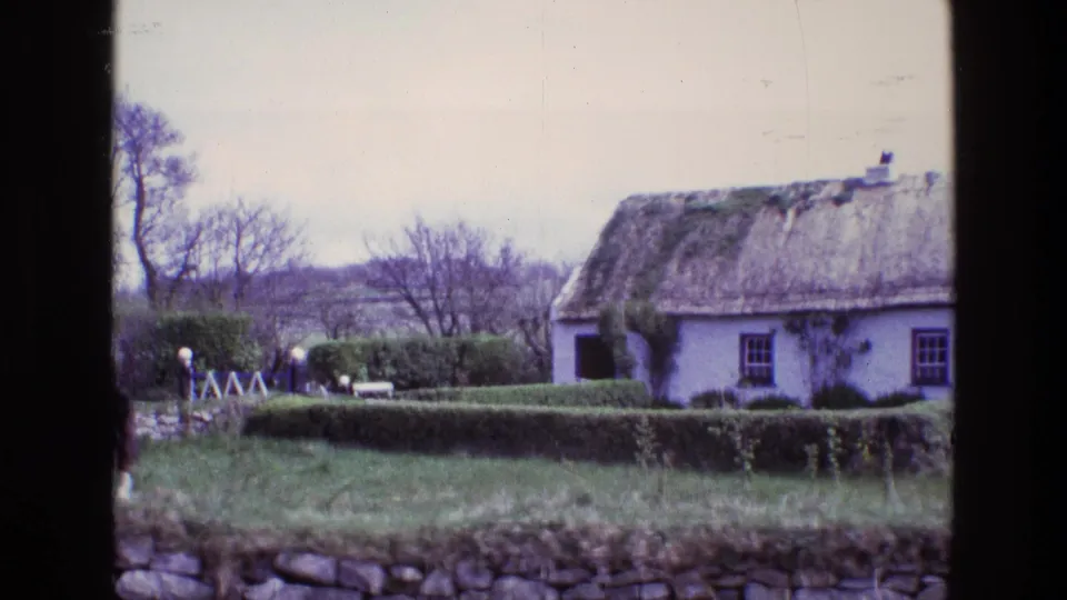 A 1969 Ireland home-movie frame centers the thatched cottage, clipped hedges, grassy yard, stone wall and small distant figures near a gate.