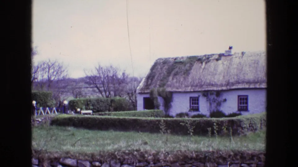 The final 1969 Ireland frame holds on the white thatched cottage, hedge boundary, grassy foreground, stone wall and distant rural fields.
