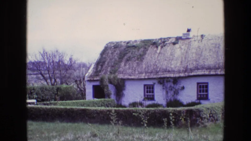 1969 Ireland 8mm frame showing the cottage closer in, with its thatch, whitewashed wall, open doorway, hedge, and low wall.