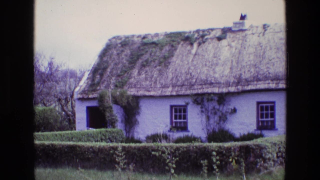 1969 Ireland home-movie frame of a whitewashed thatched cottage behind a clipped hedge, with small dark-framed windows and an open doorway.