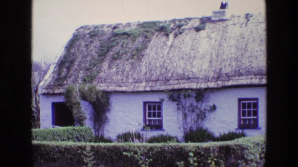 1969 Ireland view of a traditional thatched cottage with white walls, dark window frames, clipped hedge, and weathered mossy roof areas.