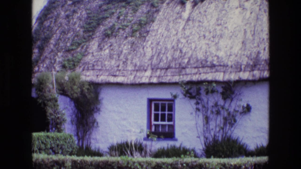 1969 Ireland home-movie frame showing a whitewashed thatched cottage, blue-framed window, clipped hedge, and shrubs.
