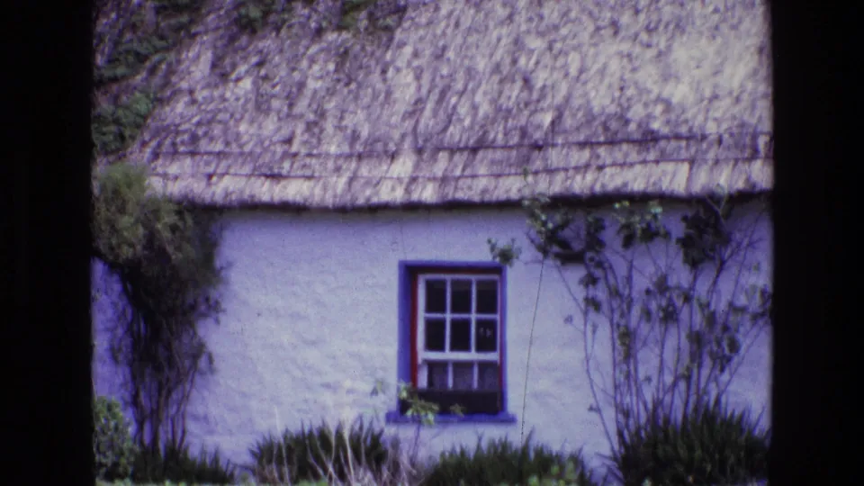 Closer 1969 Ireland home-movie view of a blue-framed divided window in a whitewashed cottage wall beneath thatch.