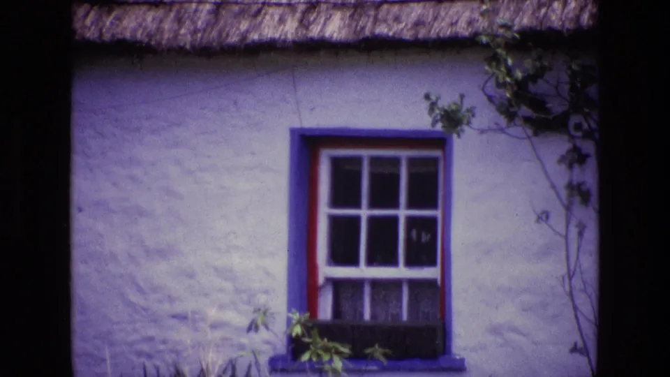 1969 Ireland home-movie frame of a thatched cottage facade with a blue-trimmed window, red accent trim, and climbing greenery.