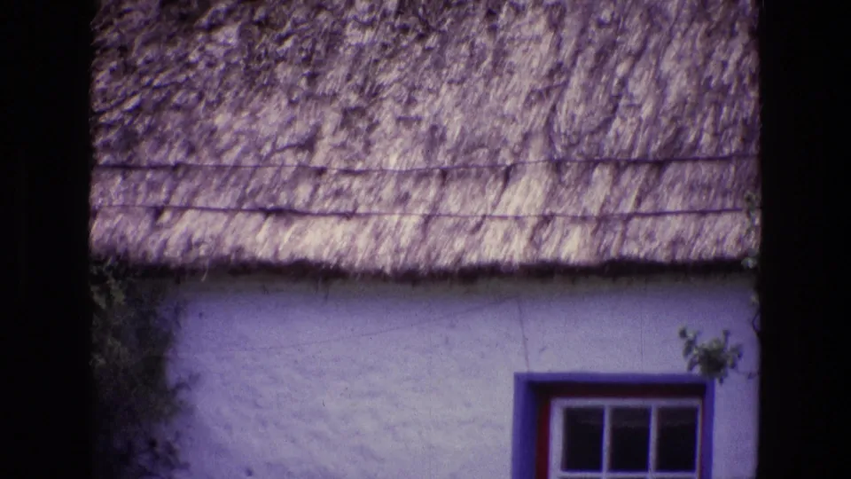 1969 Ireland home-movie frame showing more of the thatched roof and white wall, with the painted window shifted toward the lower right.