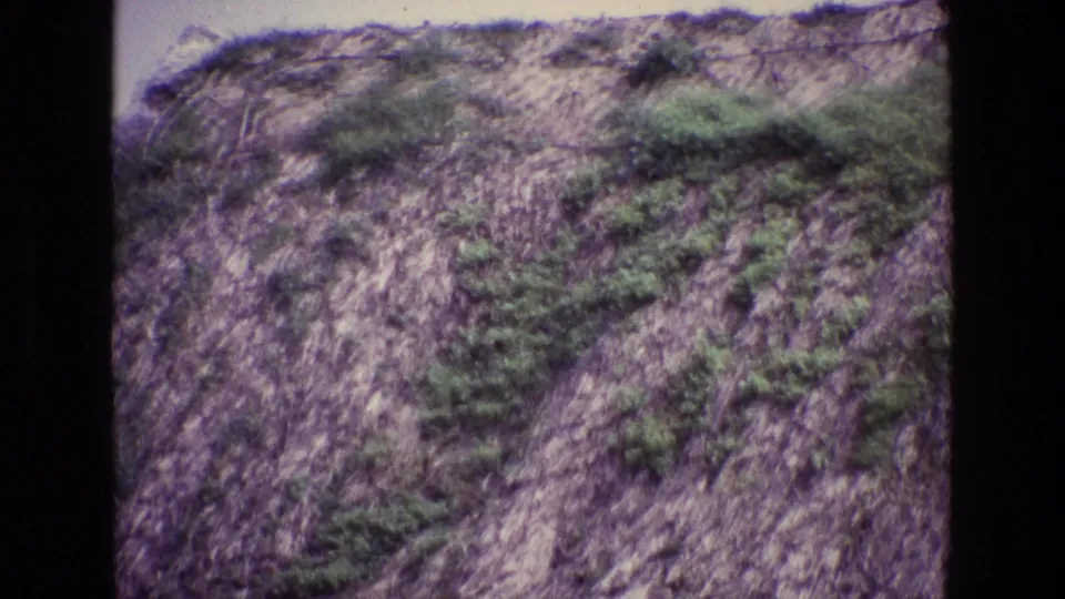 1969 Ireland home-movie frame of a steep rocky slope with scattered shrubs, faded color, and visible 8mm texture.