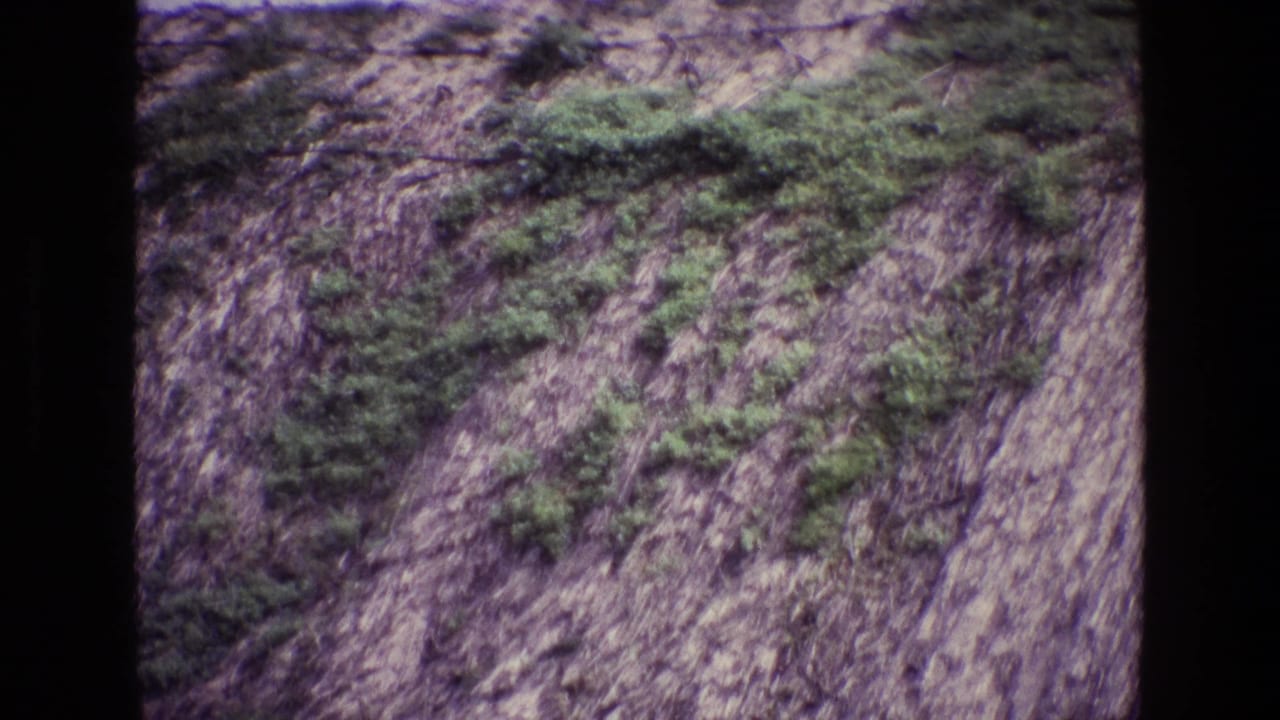 Steep rocky slope with green shrub patches and a dark vertical film border.