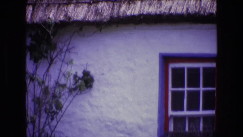 1969 Ireland 8mm frame of a white cottage exterior, thatched eaves, climbing vines, and a red and blue framed window with white panes.