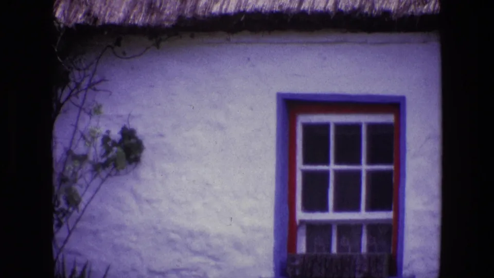 1969 Ireland 8mm frame showing a whitewashed cottage wall, thatched roof edge, vines, and a red and blue trimmed window with white muntins.