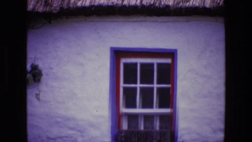 1969 Ireland 8mm frame of a white cottage wall with thatched eaves and a red and blue framed window, bordered by the dark film scan area.