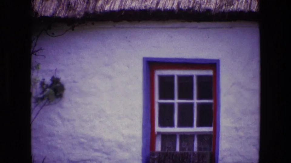 1969 Ireland home-movie frame showing a painted cottage window set in a white textured wall below thatch.