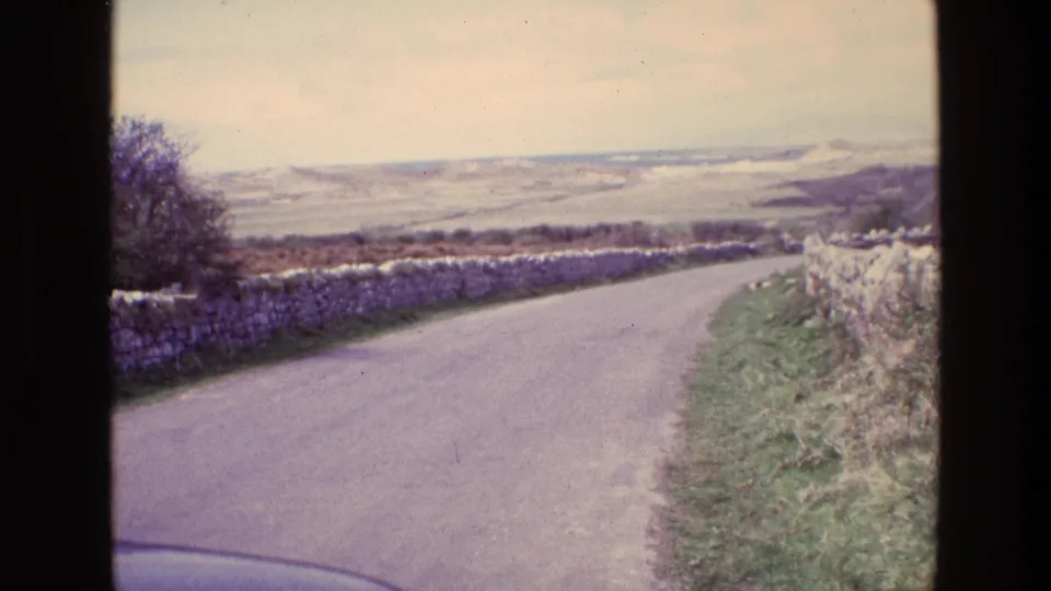 1969 Ireland home-movie frame showing a curving rural road, stone walls, grass verge, and distant hills.