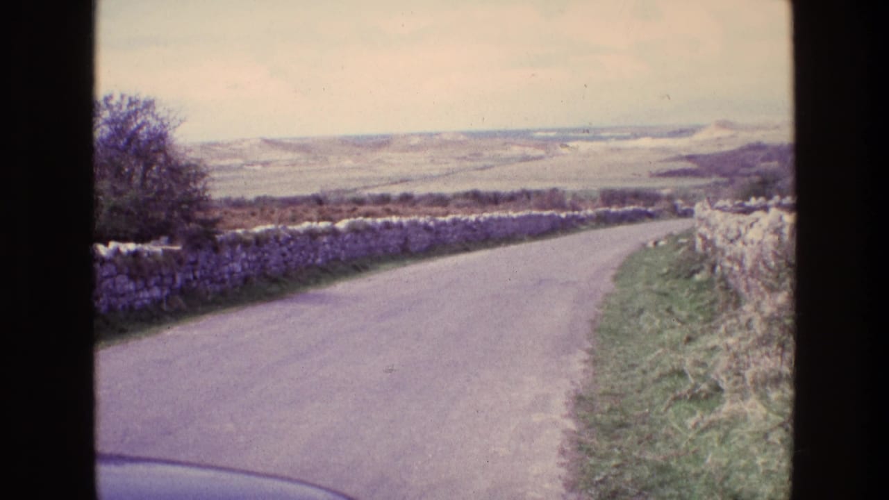 1969 Ireland 8mm frame showing a narrow rural road bordered by stone walls and grassy verges, with hills under a pale sky.