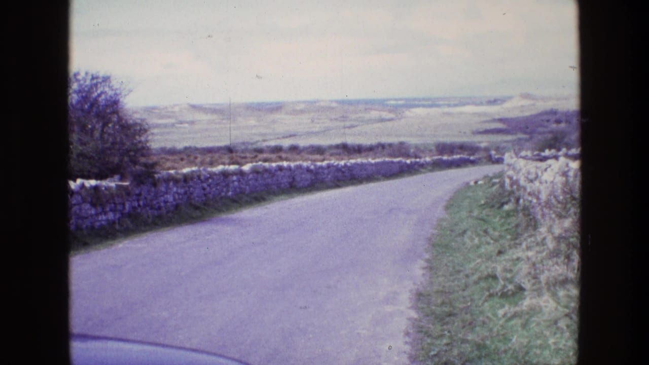 Country lane with stone walls, grass edges, and distant hills.