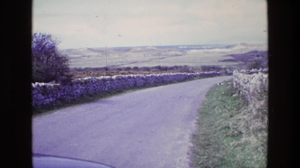 1969 Ireland 8mm frame of a quiet paved lane flanked by dry-stone walls, with open countryside stretching into the distance.