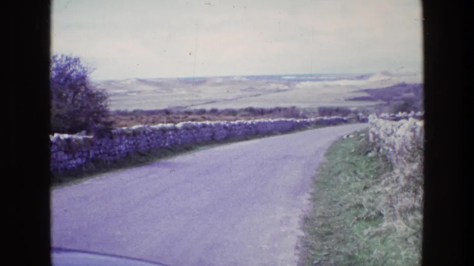 1969 Ireland home-movie frame with a rural road, stone walls, grassy banks, and soft distant hills seen through dark scan borders.