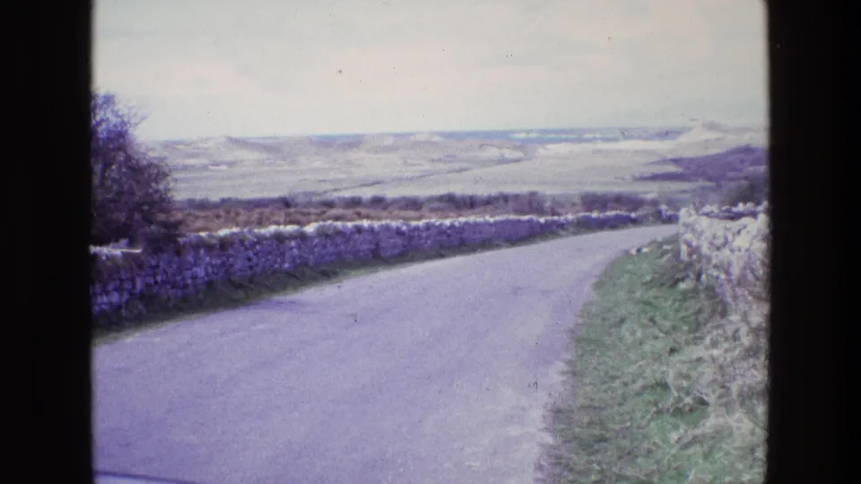 1969 Ireland 8mm frame showing a gently curving rural lane, rough stone walls, grassy roadside, and distant hills beneath a pale sky.