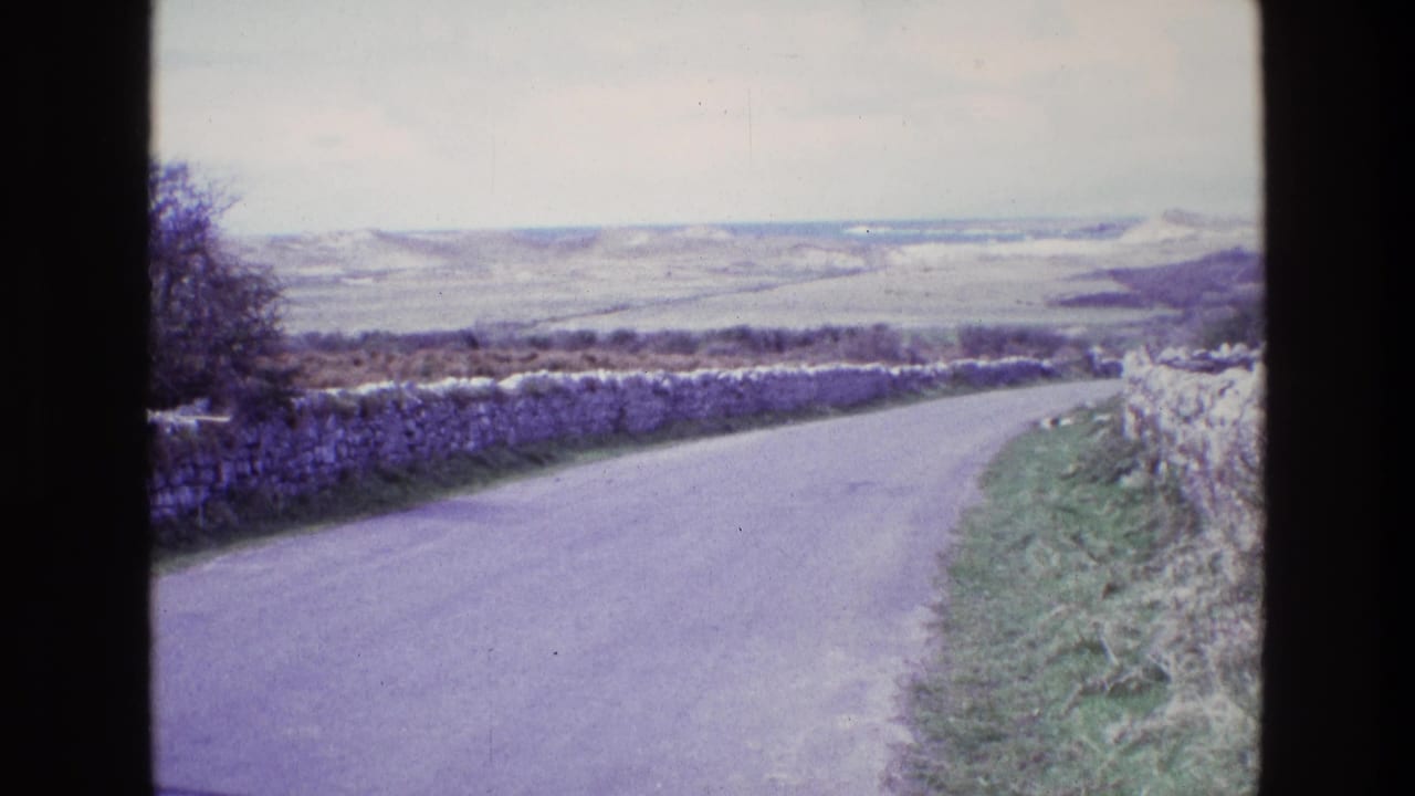Curving rural road bordered by stone walls and grassy verges under a pale sky.