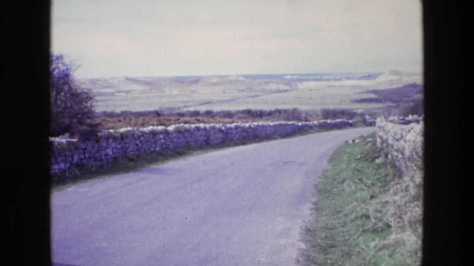 1969 Ireland home-movie frame showing a quiet rural road curving between dry-stone walls toward rolling hills.