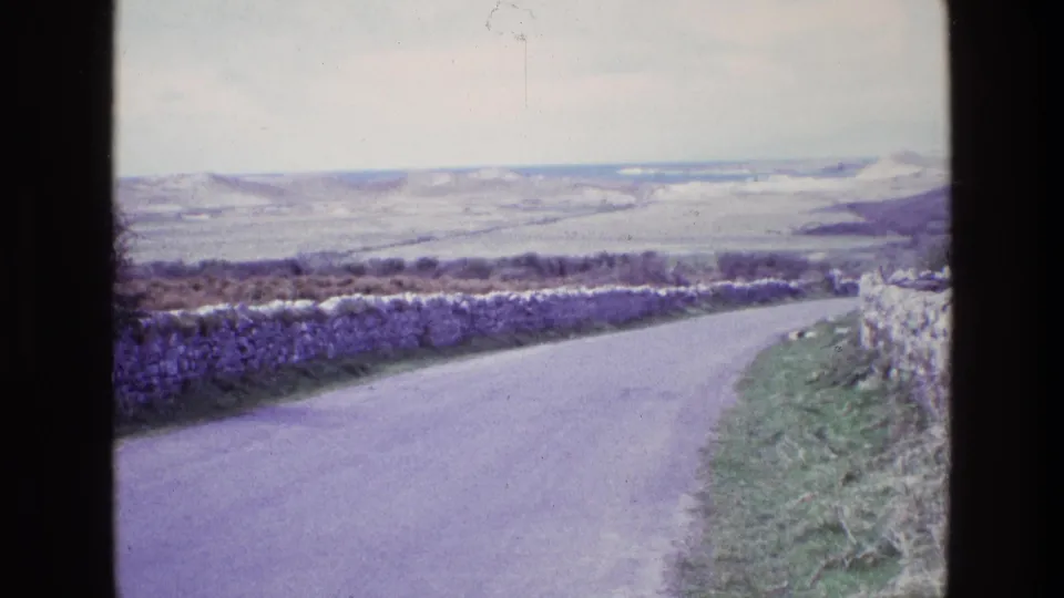 1969 Ireland home-movie still showing a rural road beside stone walls, grass, and open hillside under a pale sky.